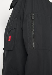 Manche de veste noire avec une poche zippée verticale et une poche poitrine avec un patch rectangulaire rouge avec un texte noir.