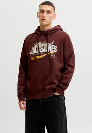 Jeune homme portant un sweat à capuche bordeaux Jack & Jones avec logo imprimé et un jean noir, debout avec une main dans la poche devant un fond uni.