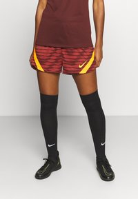 Rote und orange gemusterte Nike-Shorts mit gelben Akzenten, kombiniert mit schwarzen knielangen Socken und schwarzen Fußballschuhen mit neonfarbenen Details.