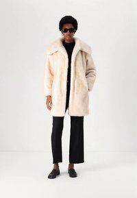Krémový kabát z umělé kožešiny se širokým límcem a volným střihem. Nošený přes černý top a černé kalhoty, doplněný černou obuví.