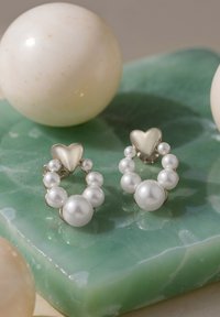 Orecchini a perno a forma di cuore in oro, decorati con perle bianche, con una finitura lucida, posati su una superficie di pietra verde chiaro.
