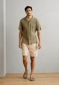 Kortærmet olivengrøn skjorte med tekstureret mønster, parret med lys beige shorts og brune sandaler, båret af en mandlig figur.