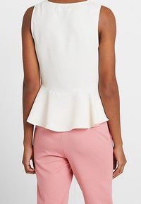 Valkoinen peplum-paita, jossa on sileä pinta, hihaton malli ja lievästi levenevä helma, yhdistetty vaaleanpunaisiin räätälöityihin housuihin.