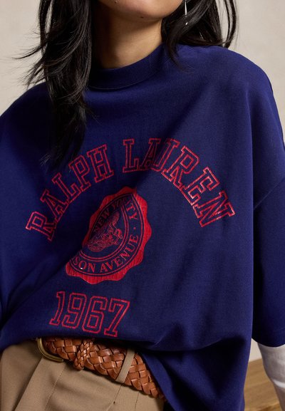 Γυναίκα φοράει μπλε φούτερ Ralph Lauren με κόκκινα γράμματα και έμβλημα, συνδυασμένο με μπεζ παντελόνια και πλεγτή καφέ ζώνη.