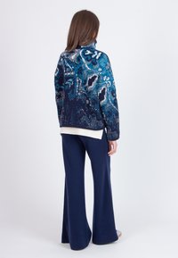 Zila un melna rakstaina adīta jaka ar augstu apkakli, kombinēta ar platām kājām zilām biksēm. Audums izceļas ar teksturētu, siltu izskatu.