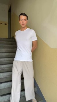 Νέος άντρας με λευκό μπλουζάκι και μπεζ παντελόνια με φαρδιά μπατζάκια που στέκεται σε εσωτερική σκάλα μπροστά σε δίχρωμο μπεζ τοίχο.