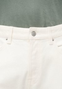 Ζώνη από λευκό τζιν με θηλιές για ζώνη και κούμπωμα με μεταλλικό κουμπί, σε συνδυασμό με πράσινο υφασμάτινο φόντο πάνω από τη μέση.