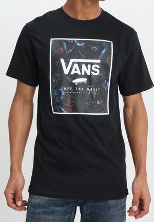 Mężczyzna nosi czarną koszulkę Vans z kolorowym tropikalnym wzorem i białym logo Vans na środku klatki piersiowej.