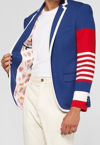 La personne porte un blazer bleu avec des bordures blanches et une manche rayée rouge, révélant la doublure de la veste à motif popcorn, assorti à un pantalon et une chemise blancs.