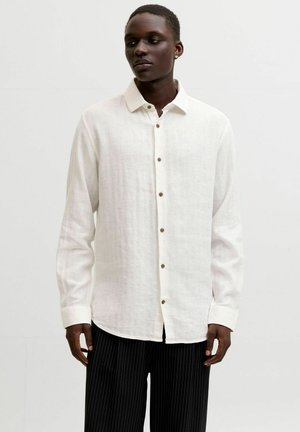 Mand iført en hvid skjorte med knapper og lange ærmer samt sorte bukser med nålestriber, der står mod en ensartet lys baggrund.