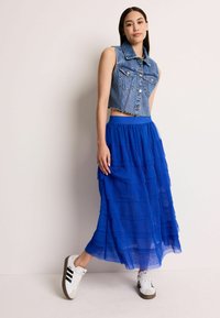 Veste en denim courte avec un ourlet effiloché, associée à une jupe maxi bleue à volants avec des plis. Des baskets blanches avec des rayures noires complètent le look.