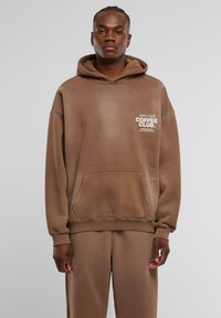 Sudadera marrón de gran tamaño con un bolsillo tipo canguro, puños acanalados y texto blanco que dice "ANOTHER COFFEE CLUB". Material suave de mezcla de algodón.