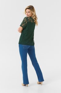 Top verde in pizzo con maniche corte, texture a pattern, abbinato a jeans svasati blu e tacchi beige. Vista posteriore che mostra la vestibilità dell'outfit.