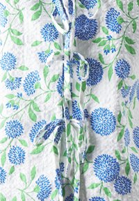 Bomullstoff med hvit bakgrunn, med blå og grønne blomstermønstre. Inkluderer knyte detalj ved halsen. Teksturert overflate.