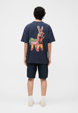 Mann mit lockigem Haar, der ein marineblaues T-Shirt mit farbenfrohem Tiermotiv und Text, gestreifte Shorts, weiße Socken und beigefarbene Sneaker trägt und steht.