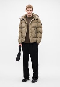 Puffervest i olivengrøn, med lynlås og trykknaplukning, hætte og sidelommer; båret over en brun sweater med sorte bukser.