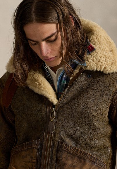 Giovane con i capelli lunghi che guarda verso il basso, indossa una giacca marrone in shearling con colletto di lana e una camicia a quadri blu sotto.