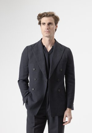Hombre con cabello castaño claro que lleva una chaqueta cruzada oscura y pantalones a juego, de pie con una mano en el bolsillo, fondo neutro.
