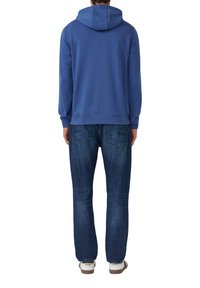 Uomo che indossa un hoodie blu, jeans blu scuro e sneakers bianche, in piedi con la schiena rivolta verso l'osservatore su uno sfondo bianco.