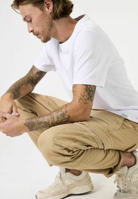 Witte katoenen t-shirt, beige cargobroek met meerdere zakken, en beige sneakers. Tattoos zichtbaar op de armen. Knielende houding.