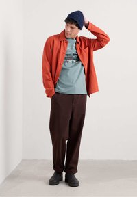 Camicia arancione a bottone, t-shirt grafica turchese, pantaloni larghi marroni, stivaletti neri alla caviglia e berretto lavorato a maglia blu navy. Sfondo semplice.