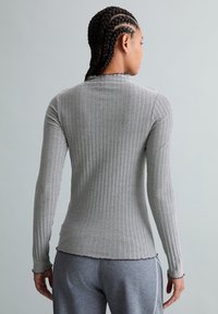 Vrouw met gevlochten haar, gekleed in een nauwsluitende, zwart-wit gestreepte top met lange mouwen en grijze broek, met de rug naar de camera tegen een effen grijze achtergrond.