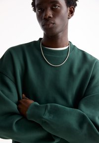 Green sweatshirt with a crew neckline, relaxed fit, and ribbed cuffs. Features a subtle logo on the sleeve and a silver chain necklace.