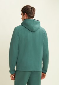 Sweatshirt à capuche en tissu texturé et doux de couleur verte. Présente une coupe décontractée avec un bord noir contrastant autour de la capuche et des épaules.