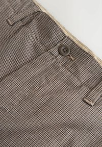 Braun kariertes Gewebe mit einer glatten Textur, versehen mit einem braunen Knopf und einem Taschendetail am Bund.