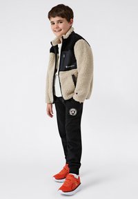 Beige en zwarte fleece jas met een rits, gecombineerd met zwarte joggingbroek met een logo patch, en feloranje sport sneakers.