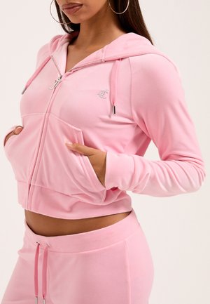 Felpa con zip in velluto rosa, dotata di tasche anteriori, cappuccio con coulisse e decorazione sul petto. Il tessuto ha una consistenza liscia.