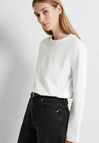 T-shirt bianco a maniche lunghe in cotone con collo rotondo, abbinato a jeans in denim neri con una fibbia decorata in oro e tasche frontali.