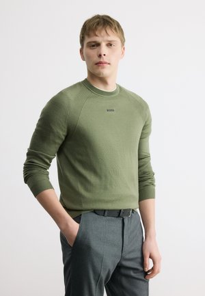 Jeune homme aux cheveux blonds courts portant un pull à manches longues vert olive et un pantalon gris, debout avec une main dans la poche.