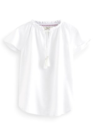 Blouse en lin blanche à manches courtes avec col à volants et lien devant avec petits pompons, présentée sur fond blanc.