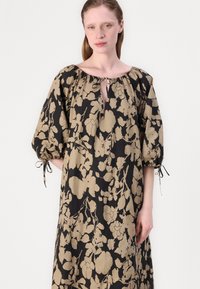 Robe noire avec un motif floral beige, coupe ample, manches bouffantes avec des liens aux poignets, et un col rond avec une ouverture en forme de clé.
