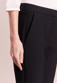 Pantalons noir ajustés avec une texture lisse, des plis subtils et une coupe slim, assortis à une blouse blanche à manches. Détails du tissu lisse visibles.