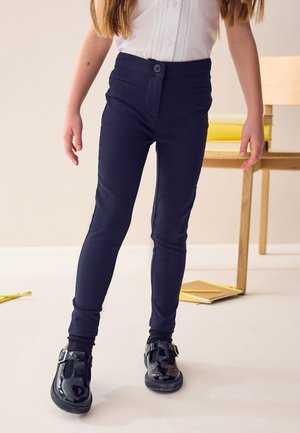Mørkeblå tætsiddende bukser lavet af strækbart stof med en frontknap, falske lommer og et elegant design, kombineret med skinnende sorte sko.