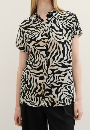 Femme portant une chemise à manches courtes noire et blanche à motifs zèbre avec boutons et un pantalon noir, debout devant un fond clair uni.