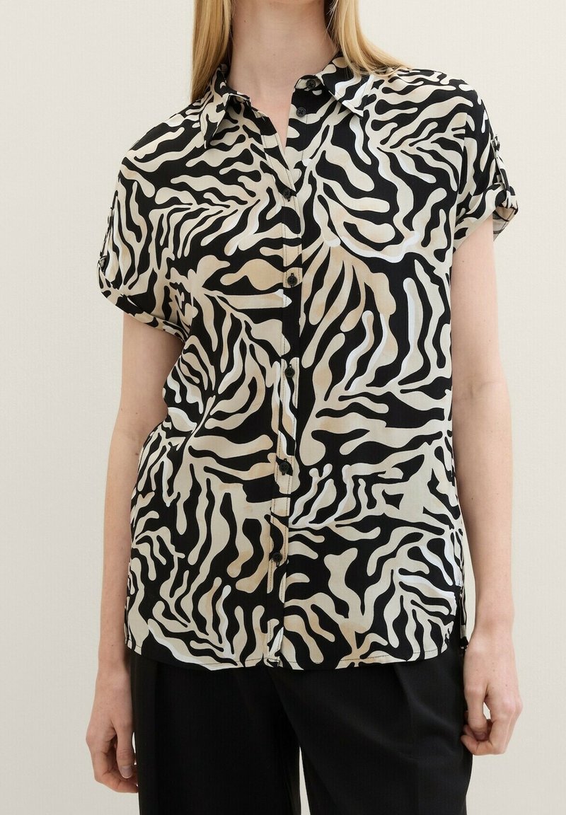 Femme portant une chemise à manches courtes noire et blanche à motifs zèbre avec boutons et un pantalon noir, debout devant un fond clair uni.