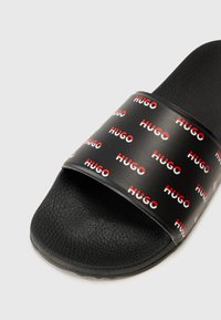 Črne natikače z zdrsno podlogo, širokim trakom in ponovljenim logotipom "HUGO" v rdeči in beli barvi na sijoči podlagi.