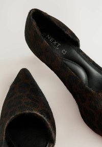 Mules à motif léopard avec un bout pointu, fabriqués en fourrure texturée et dotés d'une semelle intérieure en cuir noir. Détails de surface en peluche visibles.