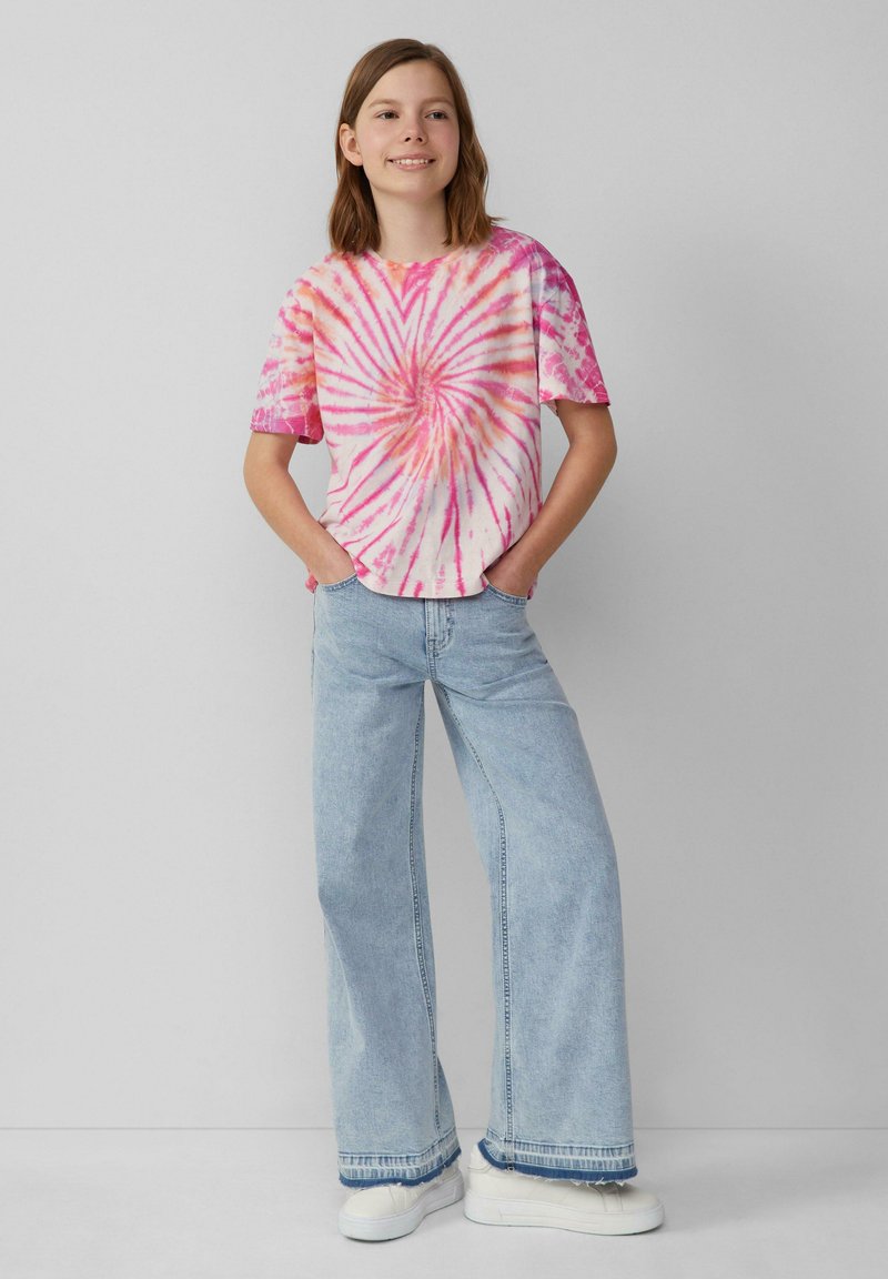 Koszulka z krótkim rękawem w kolorze różowym i białym z wzorem spirali, połączona z jasnoniebieskimi spodniami typu wide-leg i białymi trampkami.