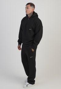 Svart zip-up hoodie gjord av mjukt tyg, i kombination med matchande svarta joggers. Båda har en logotryck med vita accenter på kläderna.