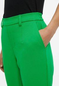 Pantalon vert sur mesure avec une texture lisse, doté de poches et d'une ceinture ajustée. La vue latérale met en évidence des plis et une coupe nette.