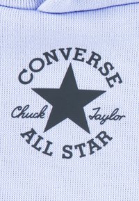 Helelillast kangast, millel on must tähelogo ja teksti "CONVERSE Chuck Taylor ALL STAR" ümmarguse mustri kujul üle tähe.