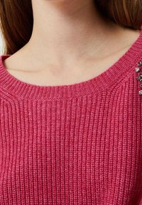 Detailní záběr na osobu oblečenou v texturovaném růžovém pleteném svetru s vroubkovaným kulatým výstřihem a dekorativními rhinestony na rameni.