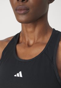 Cămașă sport neagră fără mâneci, cu decolteu rotund, evidențiind un accent cu logo alb pe partea stângă și un material neted, texturat.