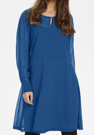 Femme portant une robe bleue semi-transparente à manches longues, longueur au genou, avec un décolleté en forme de goutte et une fermeture à boutons.