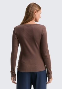 Brauner Langarmoberteil mit geripptem Muster und weitem Ausschnitt, kombiniert mit dunkelblauen Hosen. Einfaches Design mit minimalen Details.