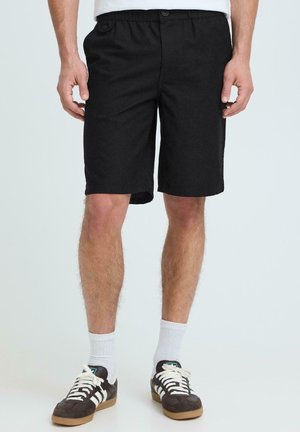 Homme portant un short noir jusqu'aux genoux, des chaussettes blanches et des baskets noires à rayures blanches, debout devant un fond uni.